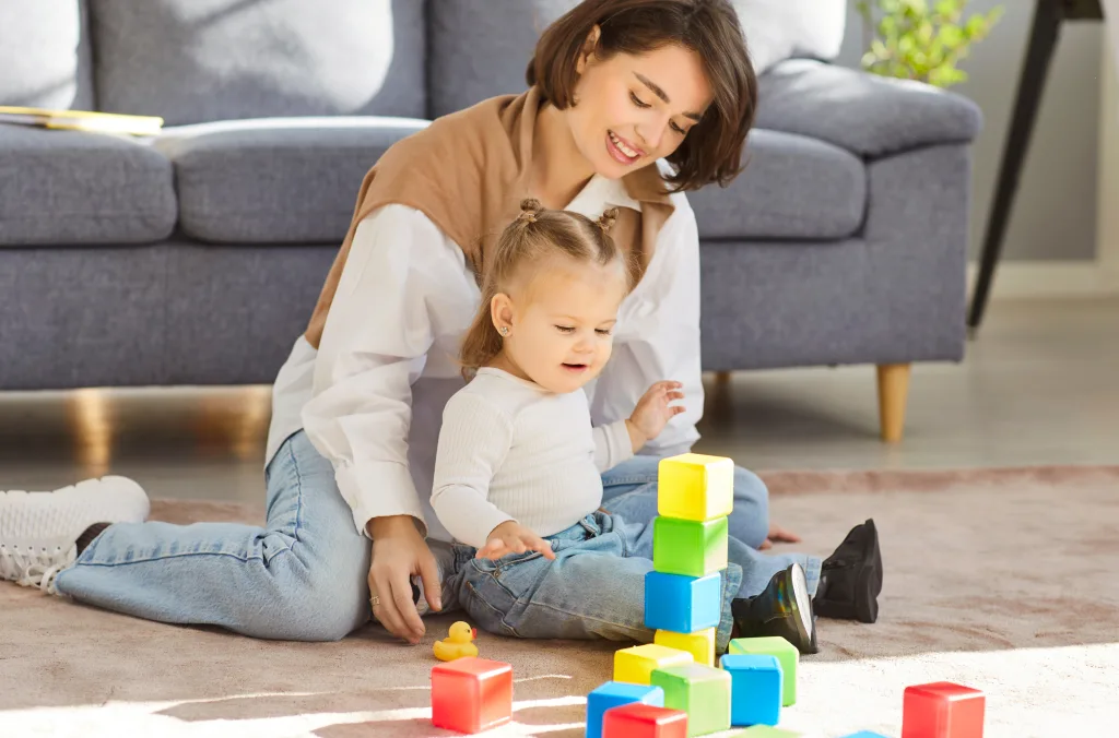 An Au Pair supports a toddler playing with colorful blocks.