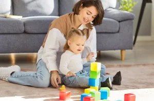 An Au Pair supports a toddler playing with colorful blocks.