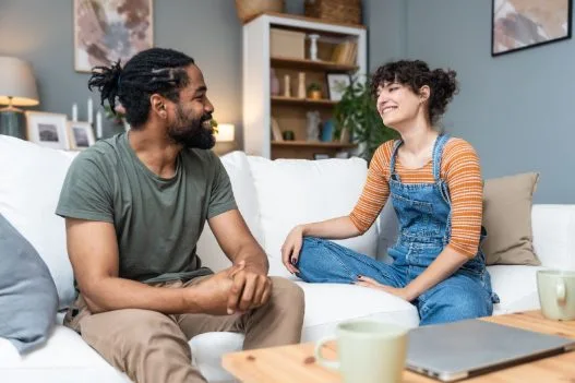 A host parent and Au Pair sit together on a couch, smiling and engaged in a relaxed, friendly conversation.