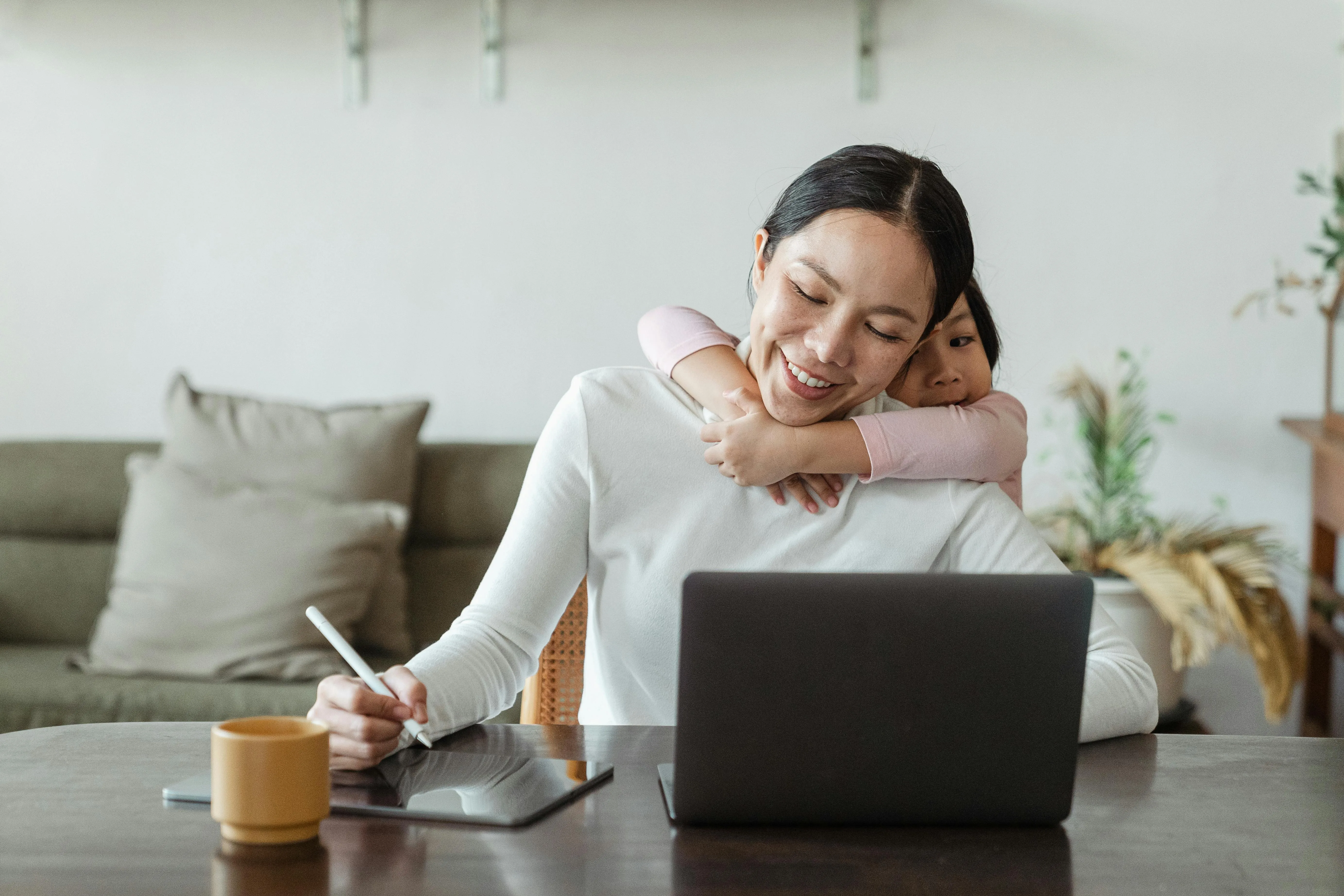 Mom working on a daily schedule on her laptop while child hugs her